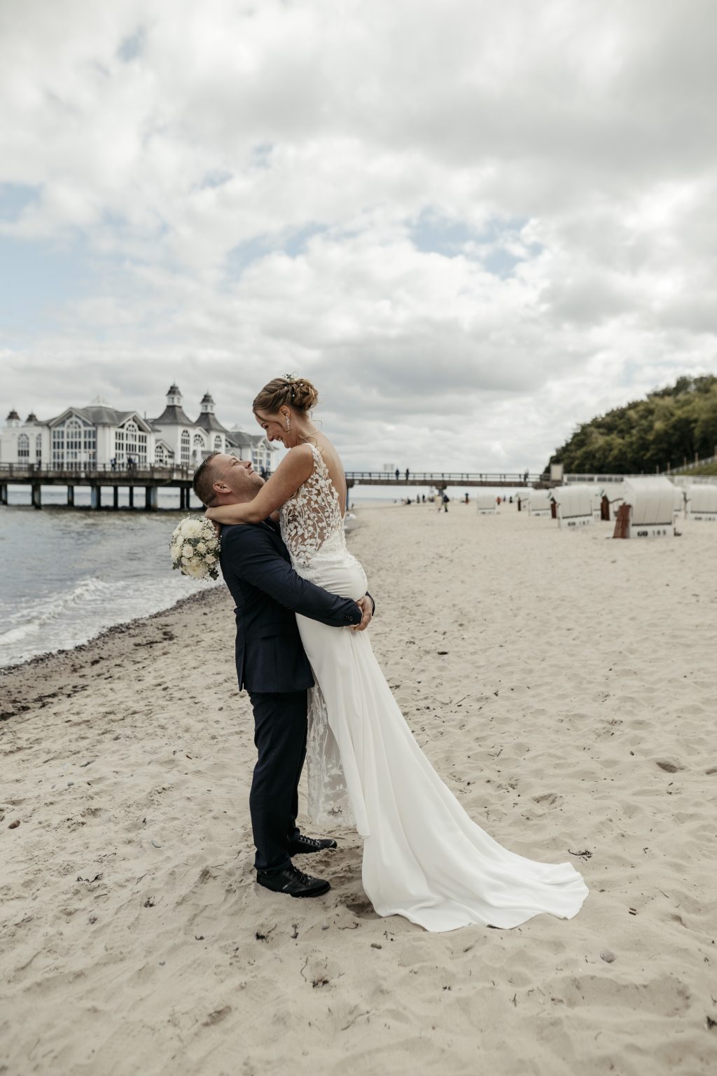 Bräutigam hebt die Braut am Strand von Sellin hoch, dahinter die Seebrücke